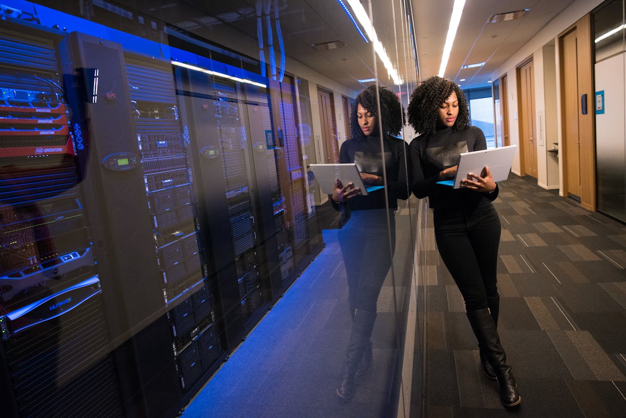Een vrouw in een zwart outfit staat tegen een spiegelwand in een datacenter, terwijl ze een laptop vasthoudt en geconcentreerd kijkt naar het scherm. Achter haar zijn servers zichtbaar in een blauw verlichte omgeving.