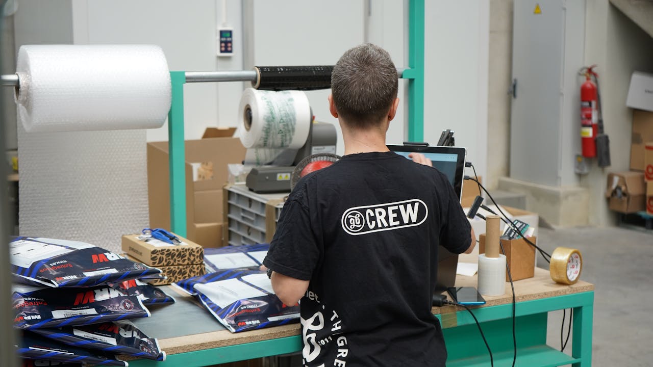 Een persoon in een zwart T-shirt werkt aan een computer te midden van verpakkingsmaterialen en dozen in een magazijnomgeving.